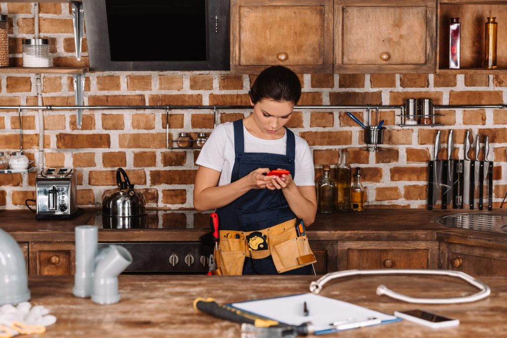 attractive young female plumber using smartphone at kitchen